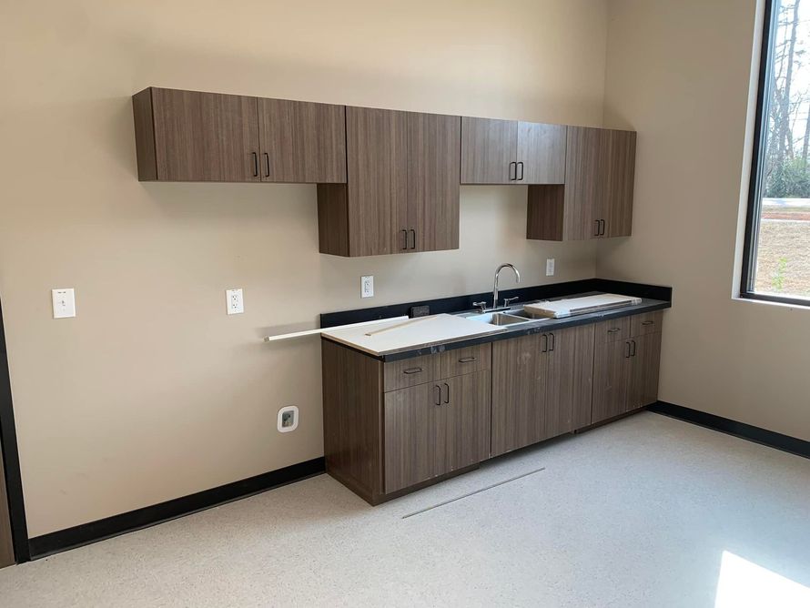 A small kitchen area with brown cabinets, a sink, and a white countertop in a room with light-colored walls and flooring.