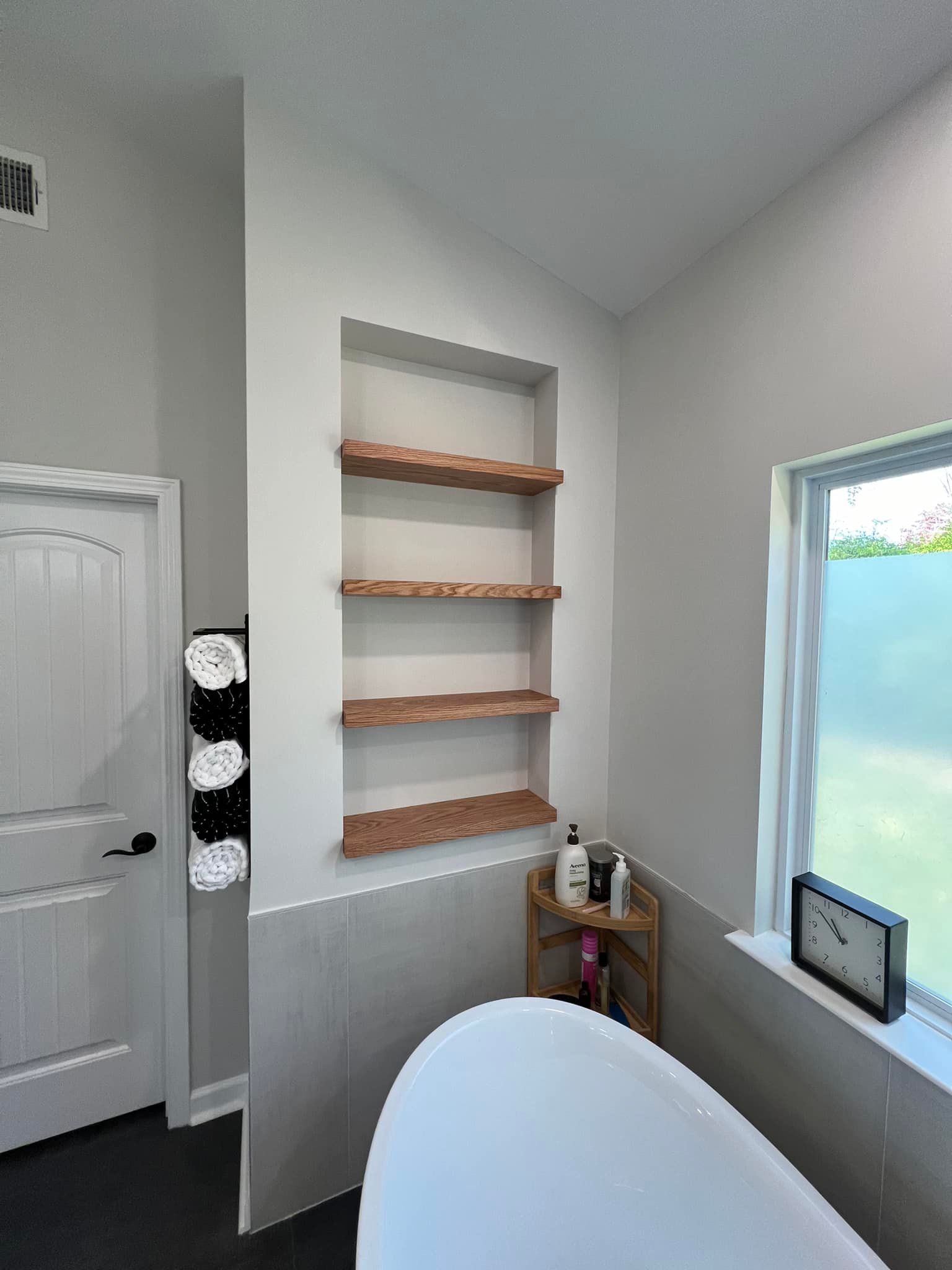 Bathroom with built-in shelves, a soaking tub, and a window. The shelves are wooden, the walls are white and gray.
