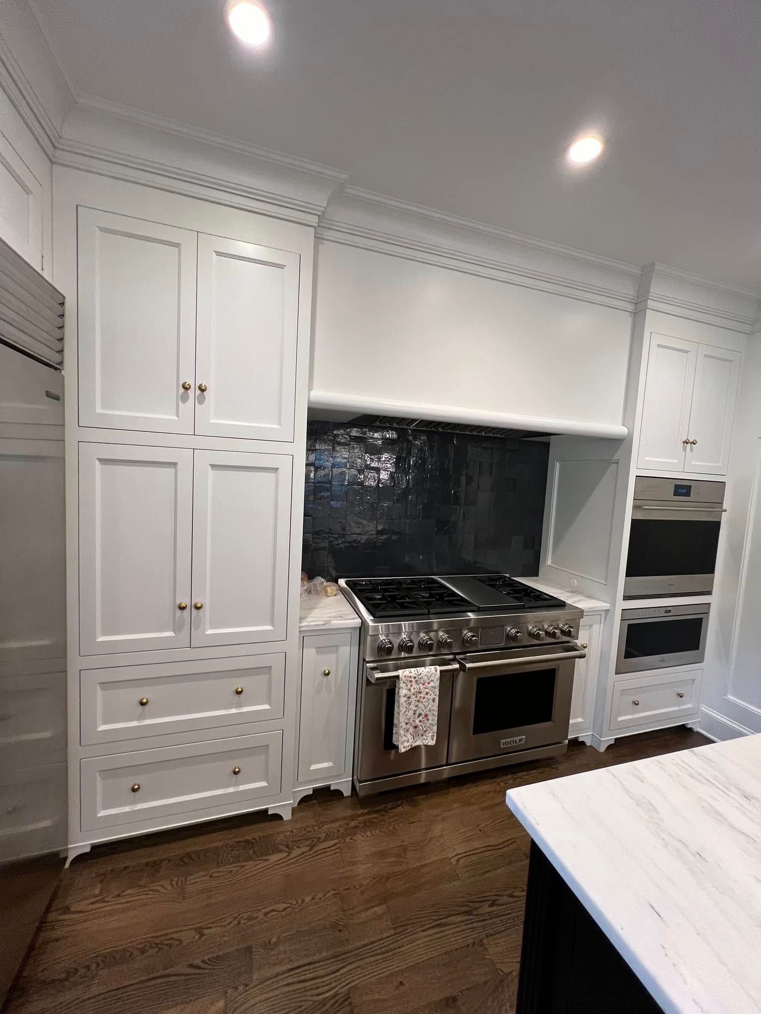 White kitchen with a stainless steel range, black tile backsplash, and built-in oven. Dark wood floors and white cabinets.