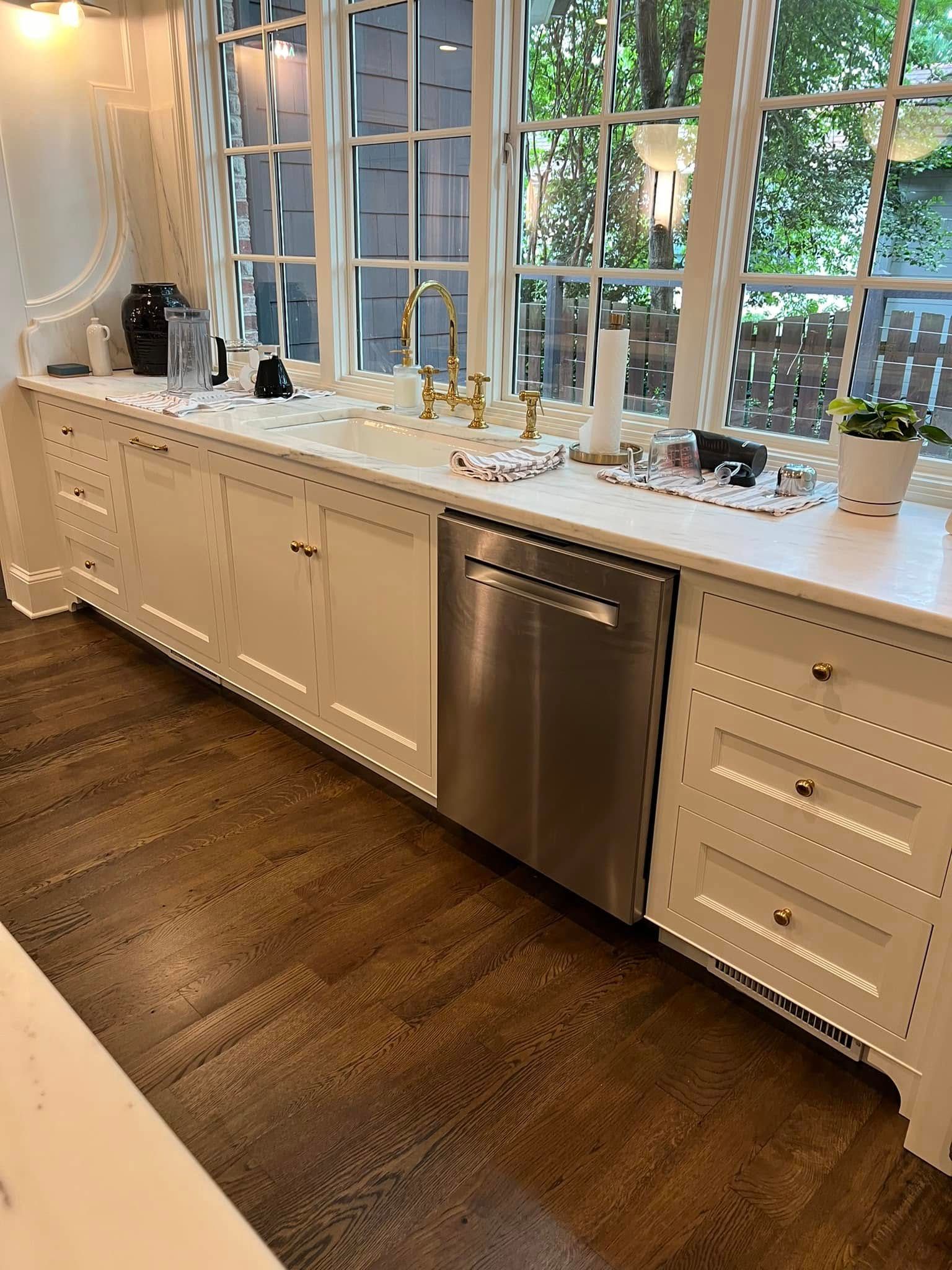 White kitchen with wooden floors and stainless steel dishwasher. Large windows provide natural light.