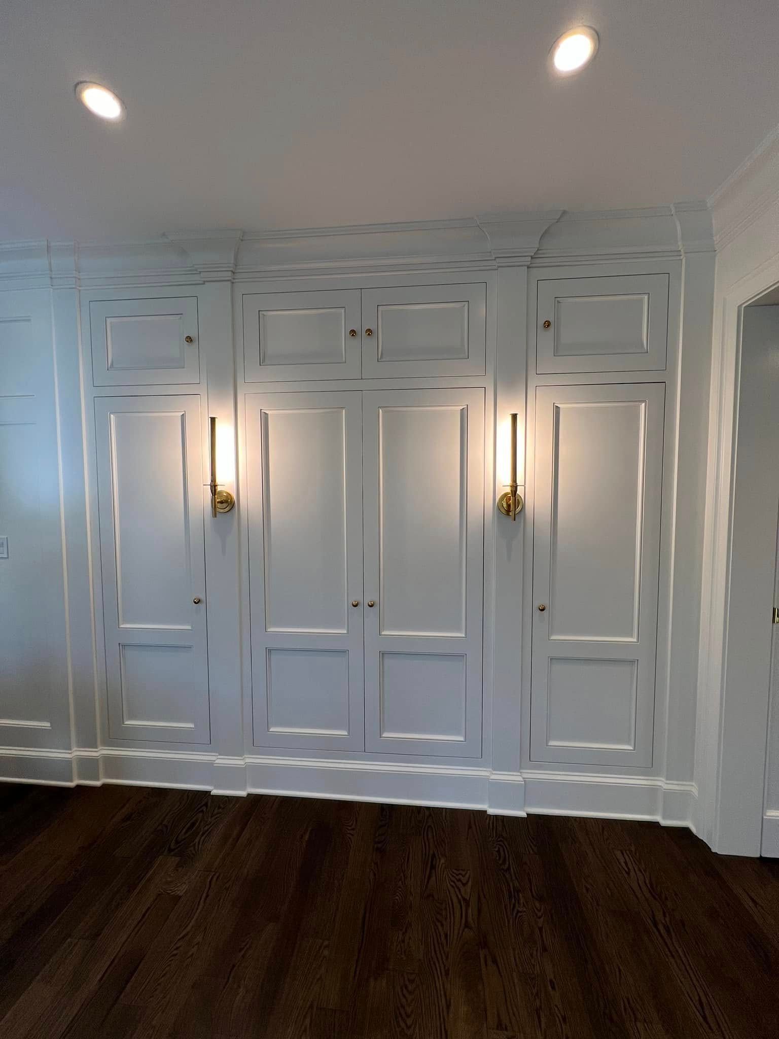 White paneled wall with built-in doors, flanked by brass sconces and lit by recessed ceiling lights; dark wood floor.