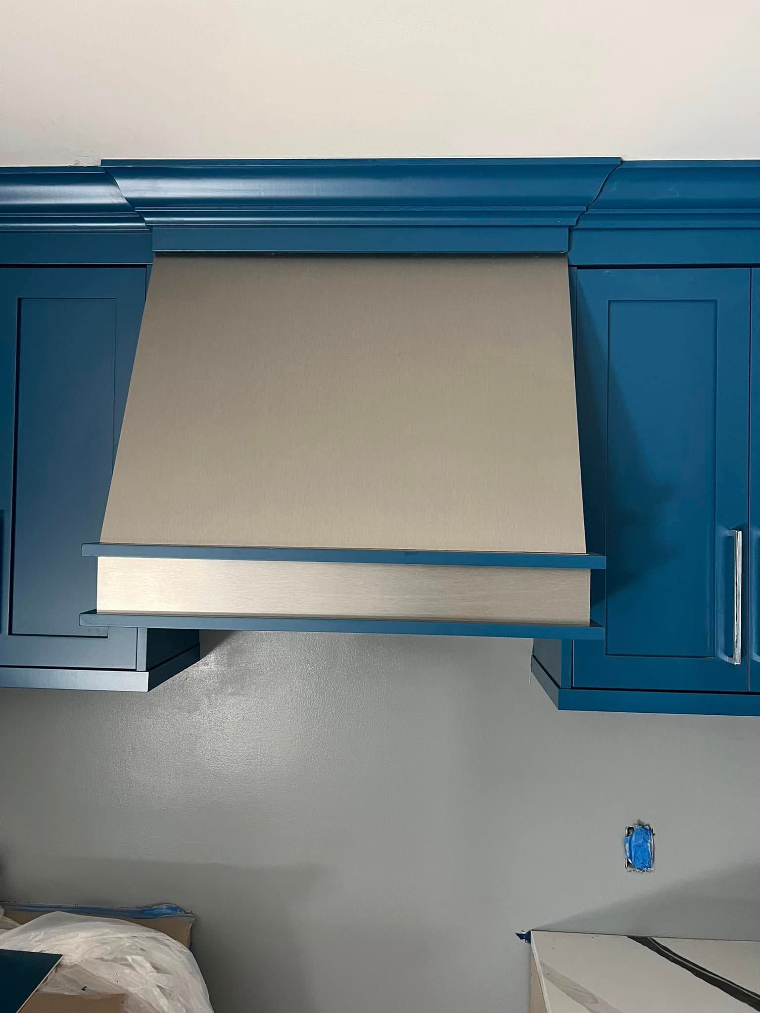 Blue kitchen cabinets with a stainless steel range hood. The cabinets are mounted above a grey wall.