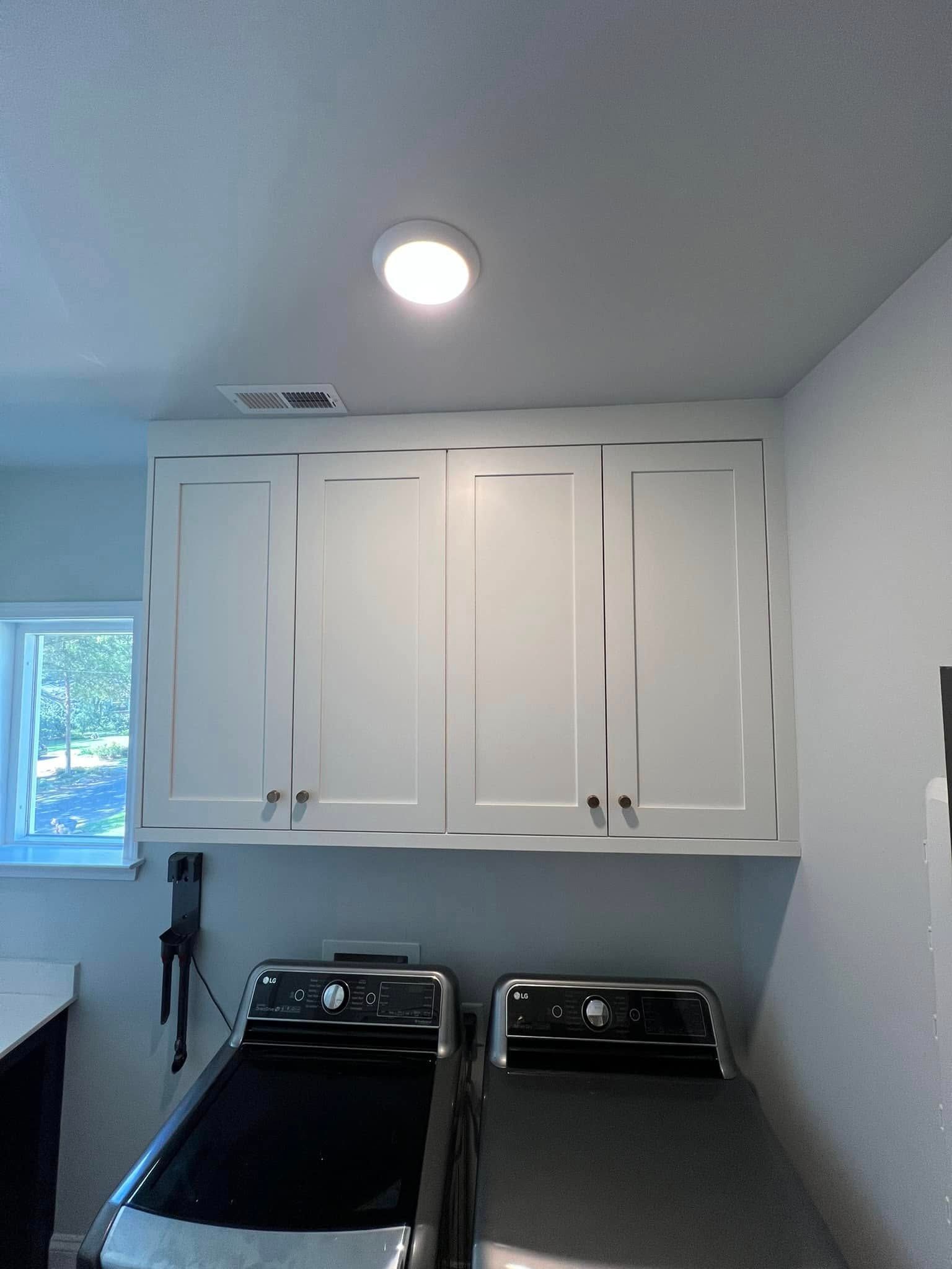 White cabinets above a washer and dryer in a laundry room, with a recessed ceiling light and a vent.