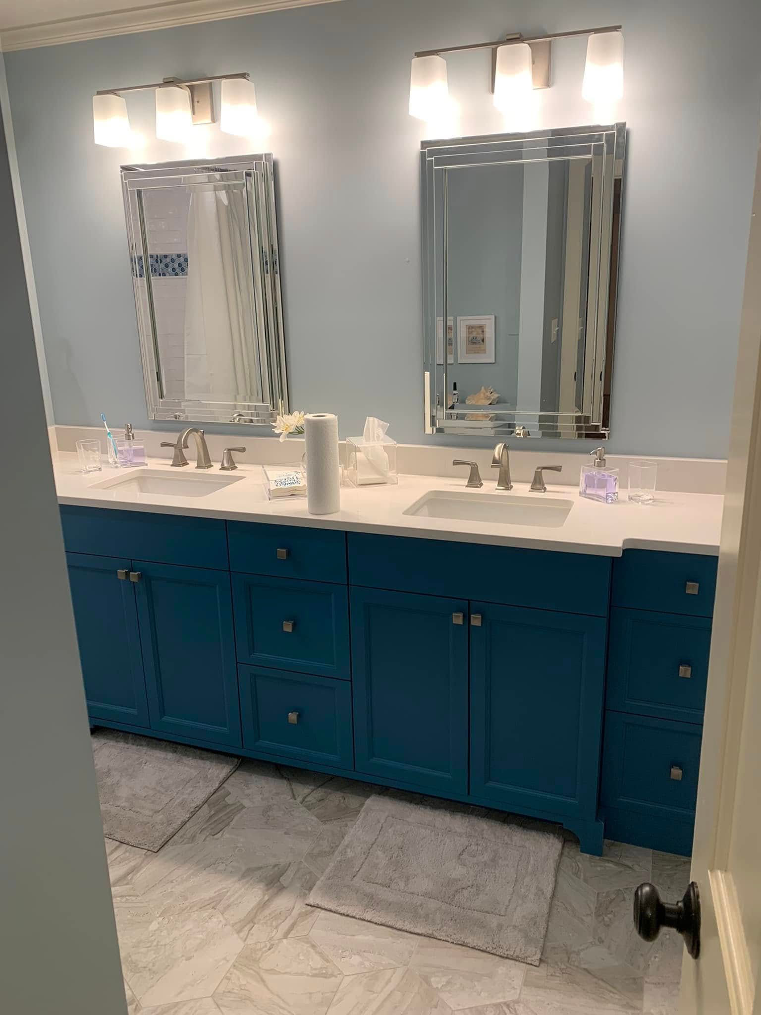 A blue bathroom vanity with two sinks and mirrors, beneath bright lights. The walls are light blue.