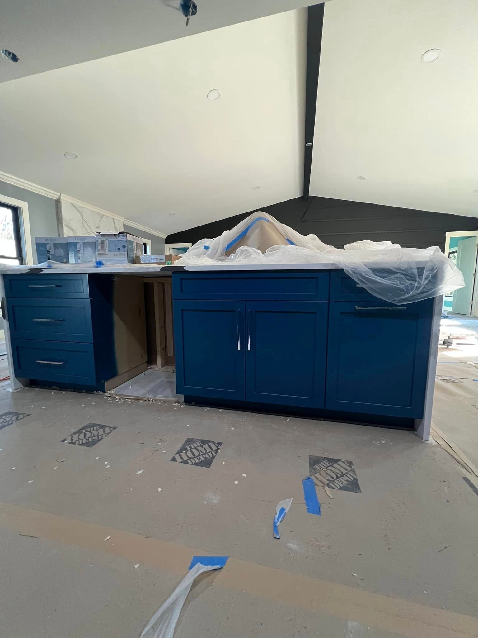 Blue kitchen cabinets are installed in a room under construction, with a white countertop.
