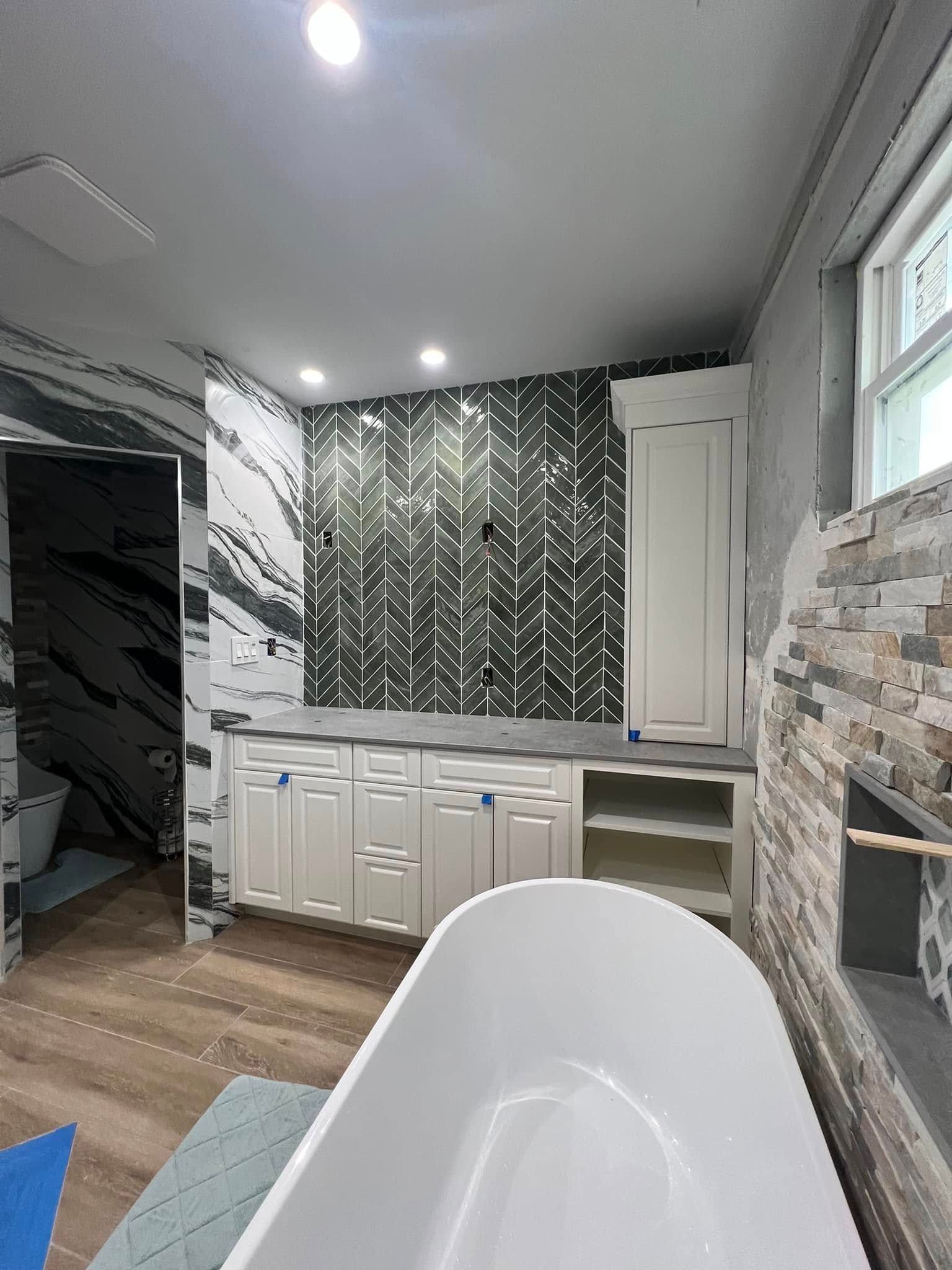 A bathroom with a white tub, white cabinets, and a dark green herringbone tile wall. A window is visible on the right.