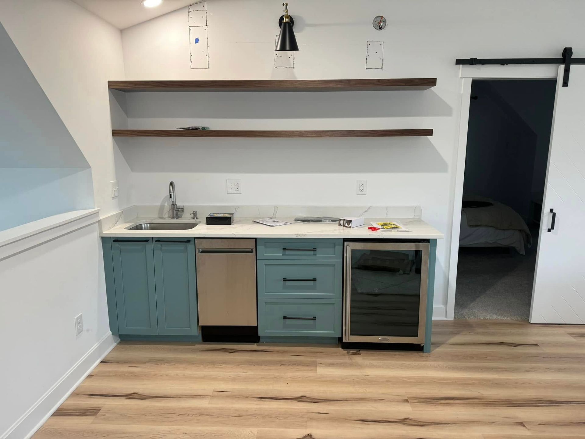 A small kitchen area with teal cabinets, a stainless steel dishwasher and wine fridge, and wooden shelves.