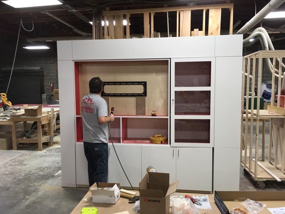 A man in a grey shirt works on a white cabinet structure with red accents. Inside a workshop setting.