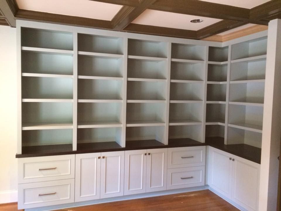 Built-in white bookshelves with lower cabinets and drawers against a wood-paneled wall in a room with a wood-beamed ceiling.