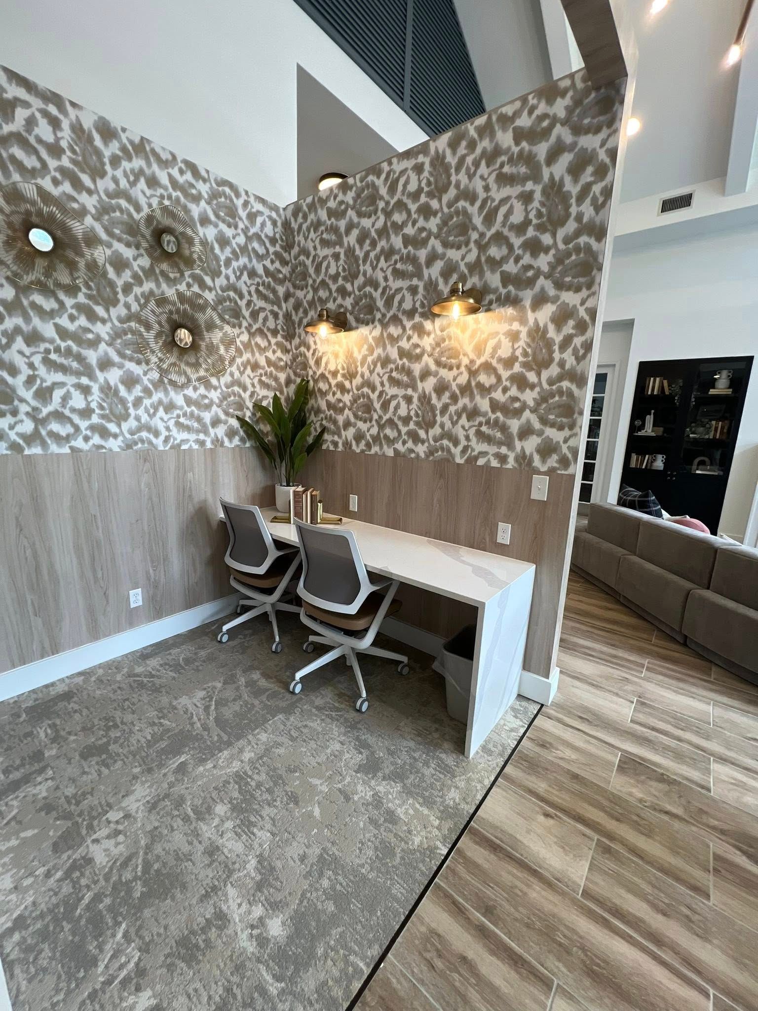 Two-person office nook with patterned wallpaper and wood paneling. Desks, chairs, plants, and sconces complete the space.