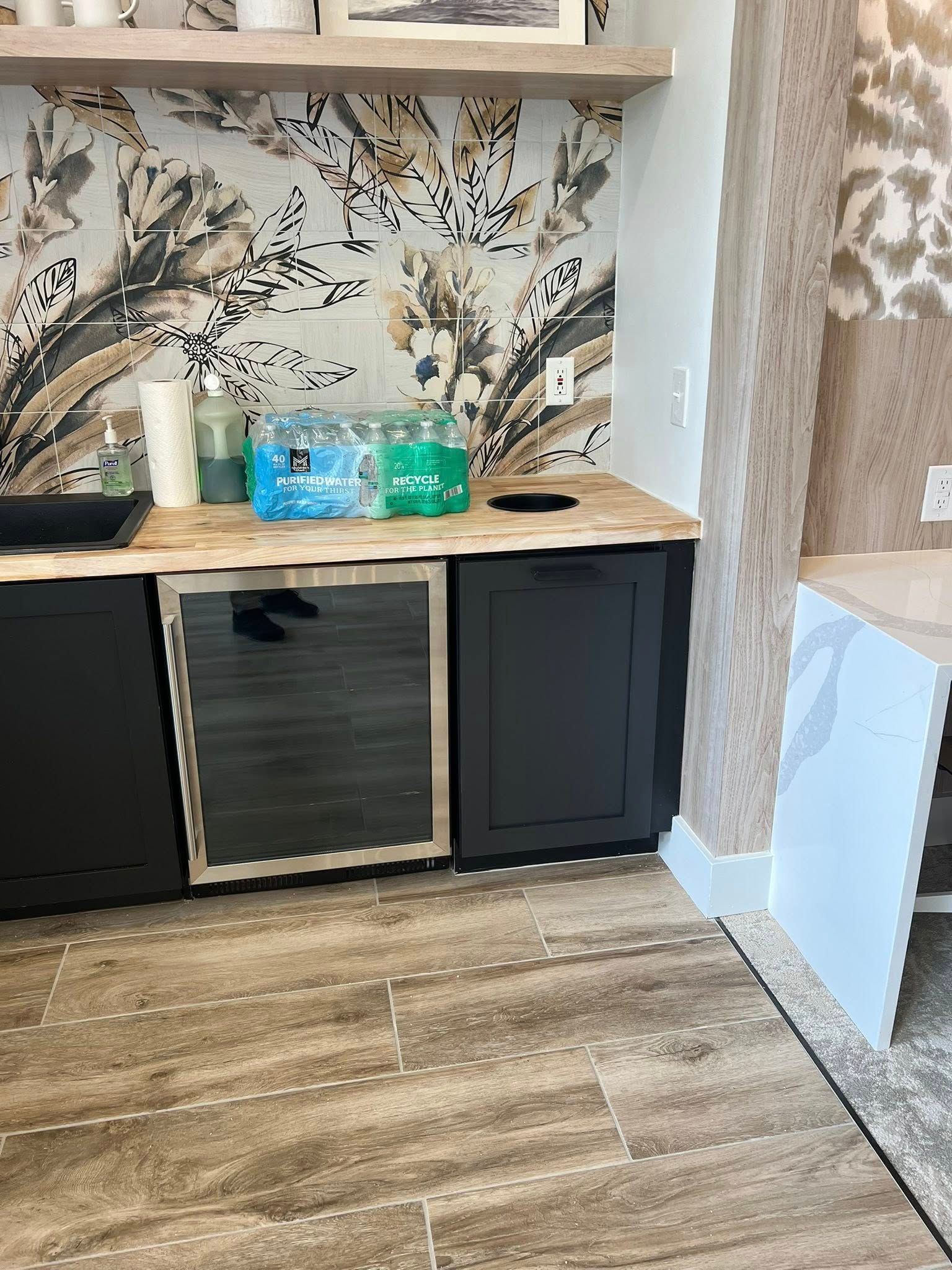 Counter with a small refrigerator, black cabinets, and jars, set against a floral wallpaper.