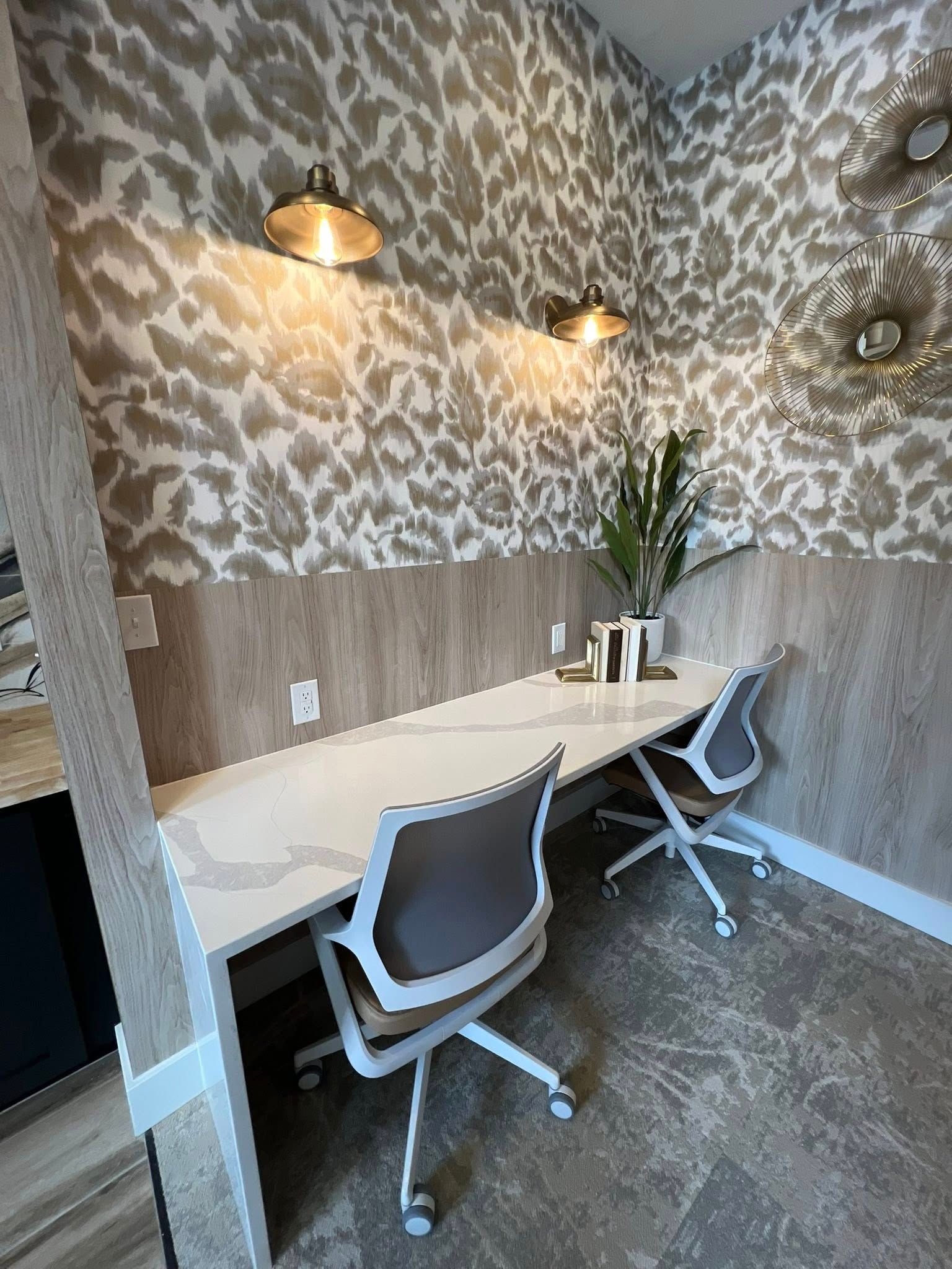Two office chairs sit at a long white desk against a wall with patterned wallpaper and sconce lighting.