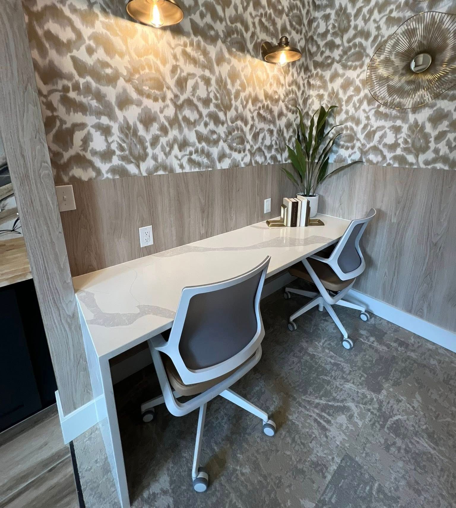 Two office chairs sit at a white desk with a patterned wall, flanked by wood paneling. A plant and books sit on the desk.