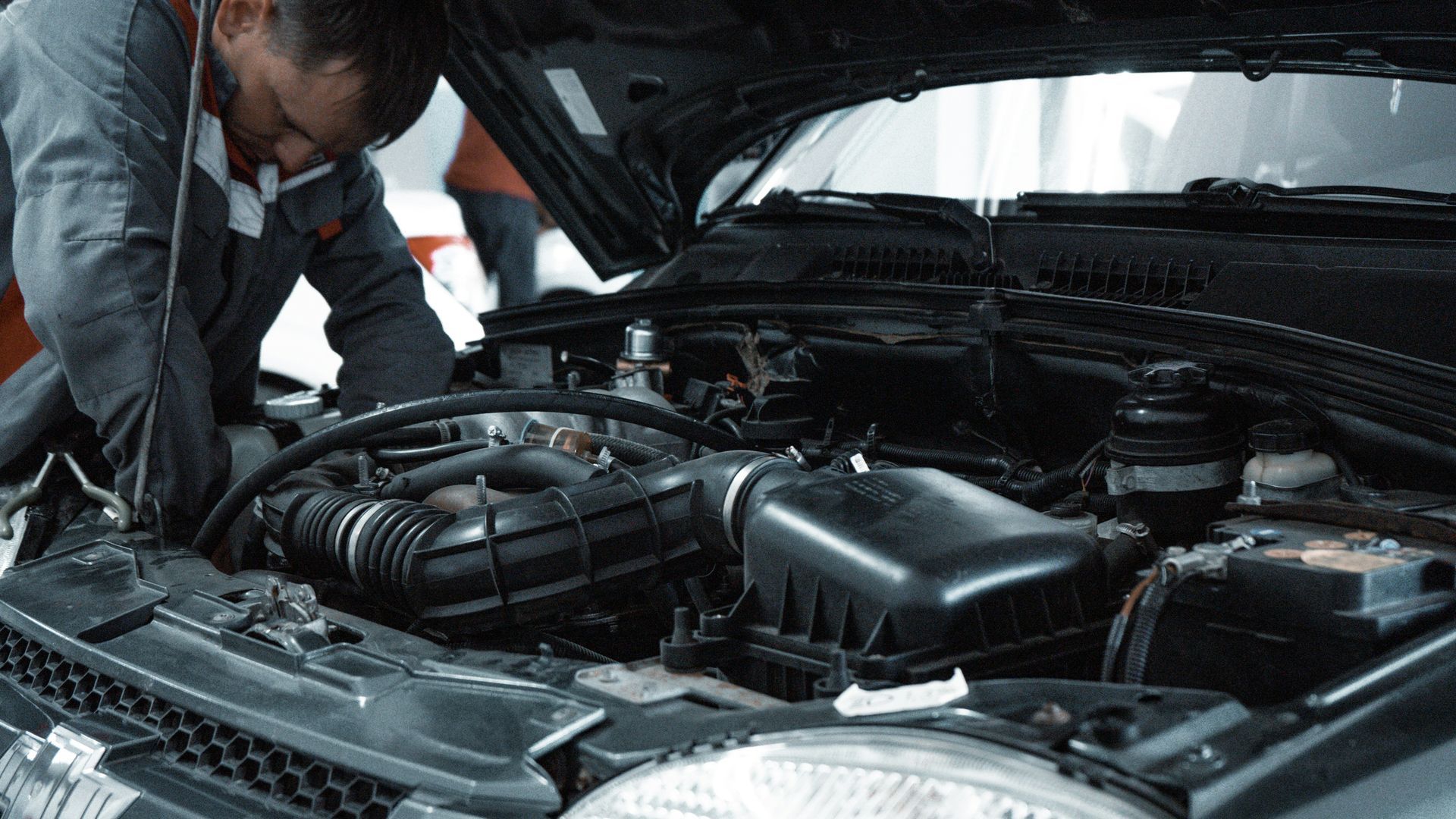 Mechanic in gray jumpsuit working on the engine of a black car with the hood open.