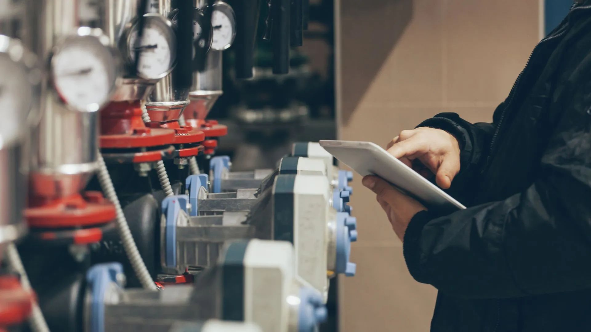 A technician uses a tablet to monitor equipment near pressure gauges and valves in a mechanical room