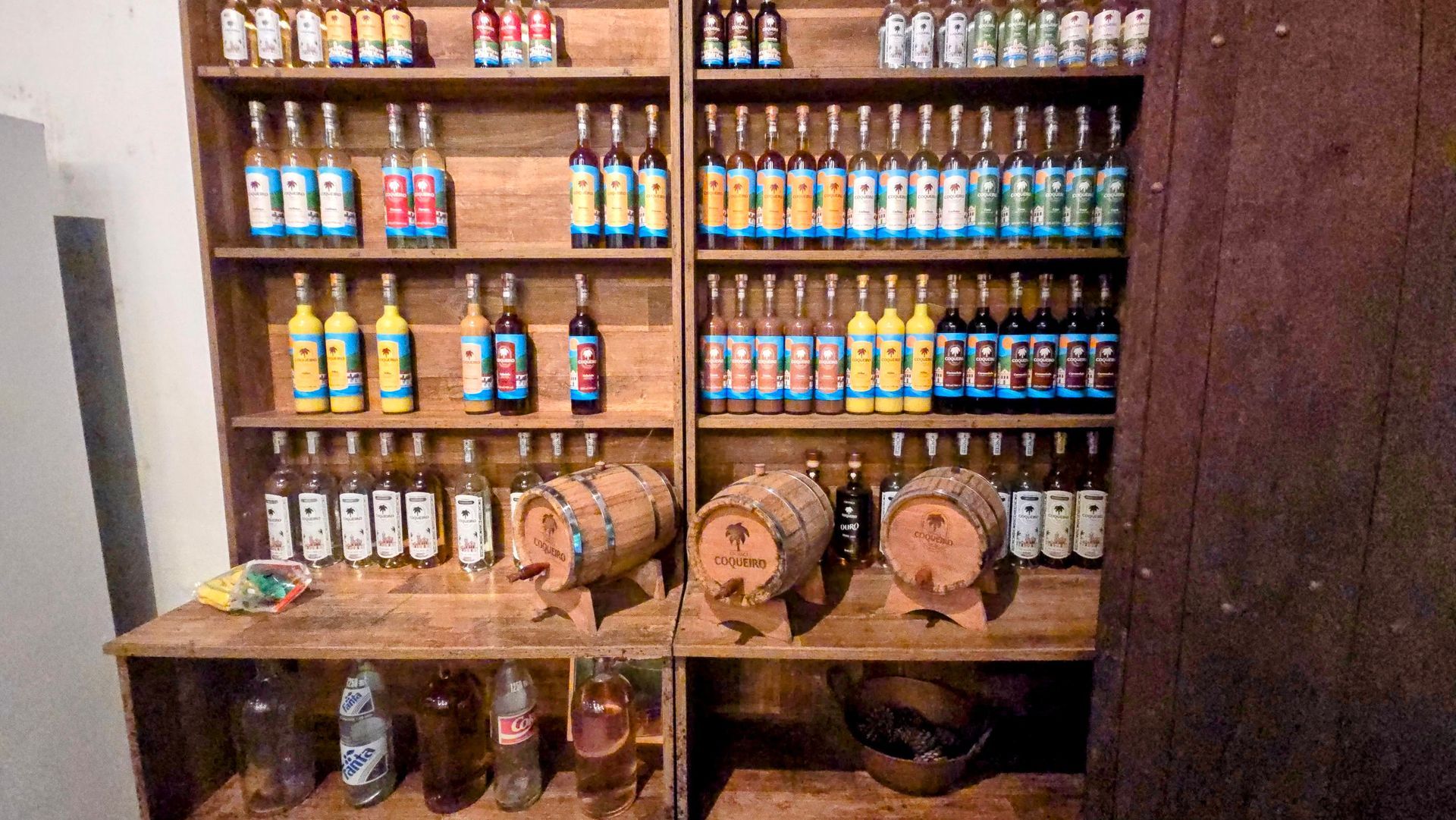 Wooden shelves displaying a variety of artisanal cachaça bottles and small wooden barrels inside a traditional distillery in Paraty, Brazil.