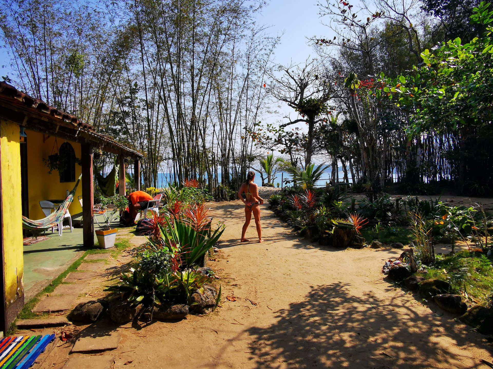 Charming outdoor area of a camping site in Ilha Grande, featuring a yellow building with hammocks, tropical plants, and bamboo trees, with a path leading to a view of the ocean, creating a relaxing and inviting atmosphere