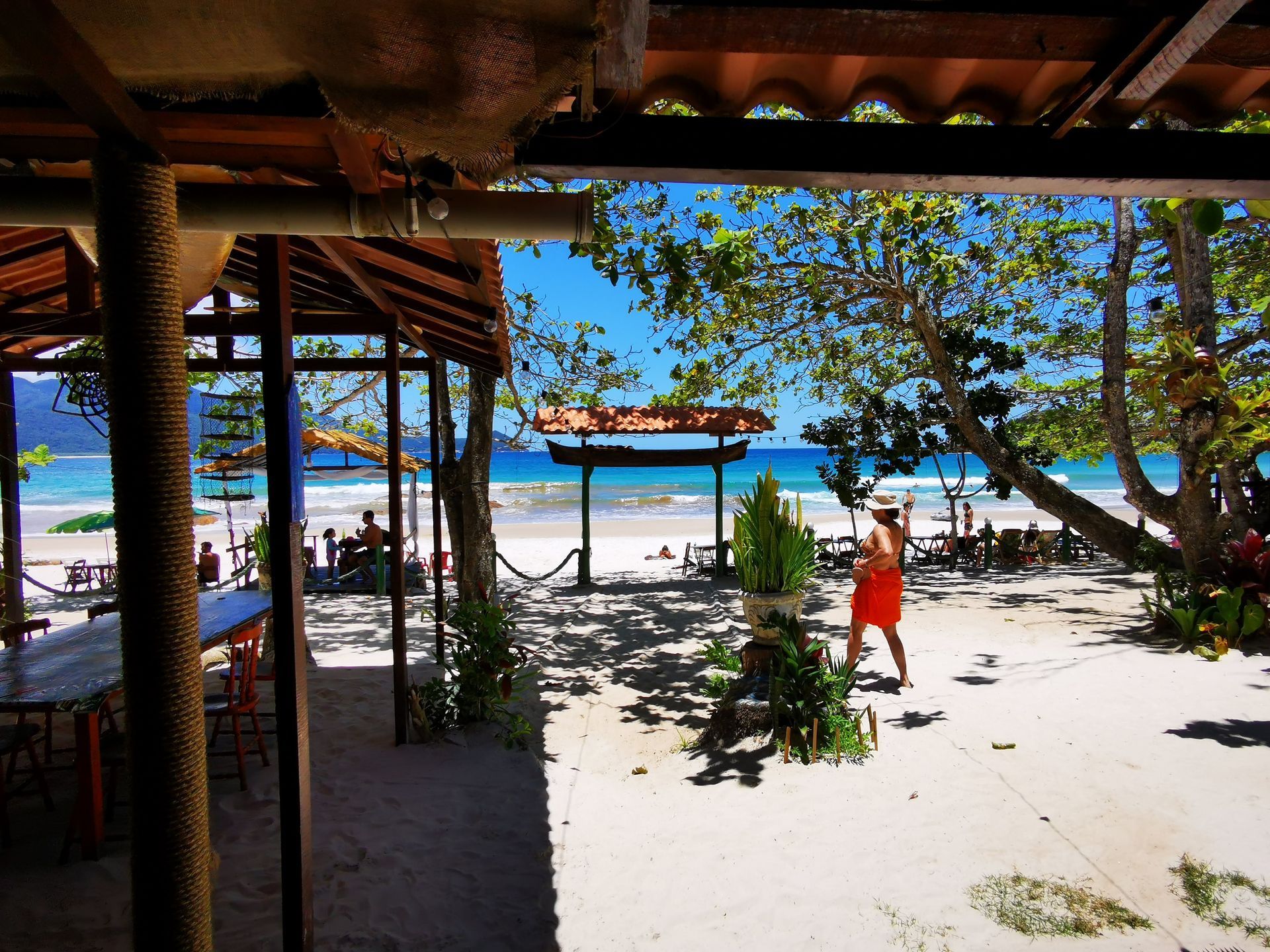 Scenic beachfront view at Aventureiro Beach, Ilha Grande, with shaded seating areas, tropical trees, and visitors enjoying the sandy shore and crystal-clear ocean, creating a vibrant and relaxing coastal atmosphere