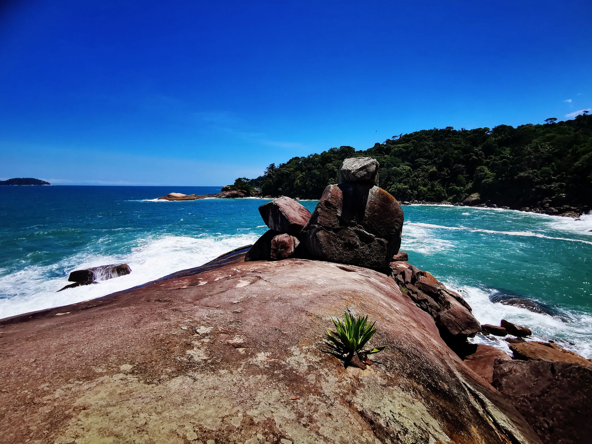 Vista panorâmica de Caxadaço, Ilha Grande, com formações rochosas imponentes, ondas do oceano turquesa quebrando na costa e uma exuberante floresta verde ao fundo, capturando a beleza selvagem e intocada desse paraíso escondido.