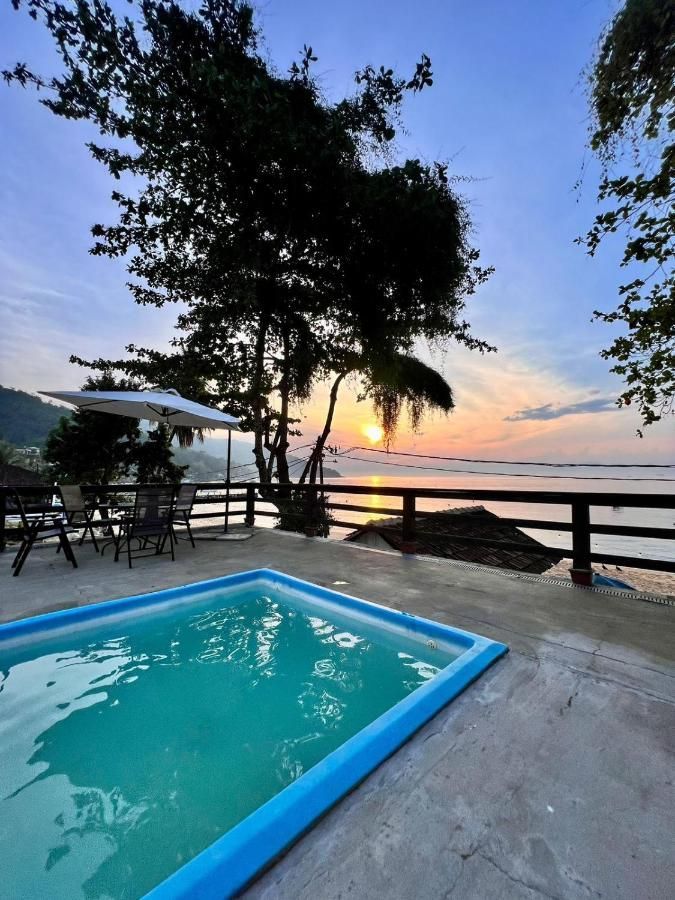 Scenic view from a pool deck at Espaço Vila Ará in Araçatiba, Ilha Grande, overlooking the ocean and surrounded by tropical trees, with a stunning sunset casting a warm glow over the water, creating a peaceful and idyllic atmosphere