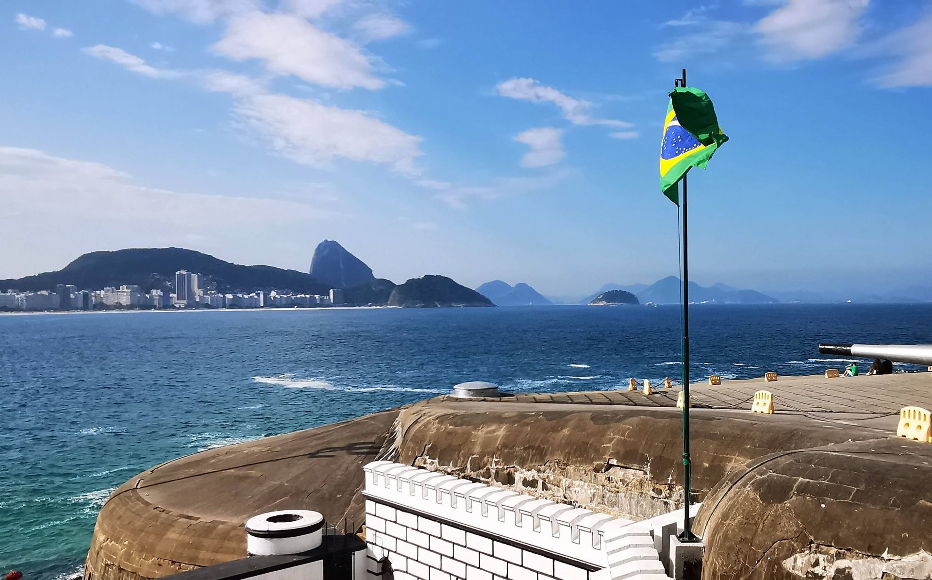 Vista do Forte de Copacabana com a bandeira do Brasil em destaque e o Pão de Açúcar ao fundo. 