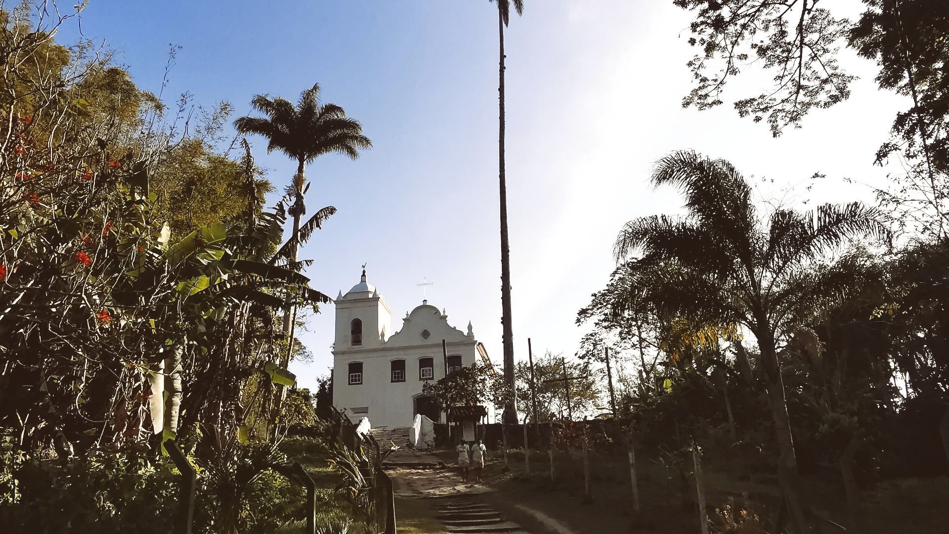 Igreja de Freguesia de Santana, Ilha Grande, Angra dos Reis, RJ, Brasil. Vista de uma trilha cercada por vegetação tropical, com palmeiras e árvores ao redor. A igreja branca com fachada simples se destaca ao fundo, com uma torre à esquerda e uma cruz no topo. O céu está claro e levemente azul, sugerindo um fim de tarde.