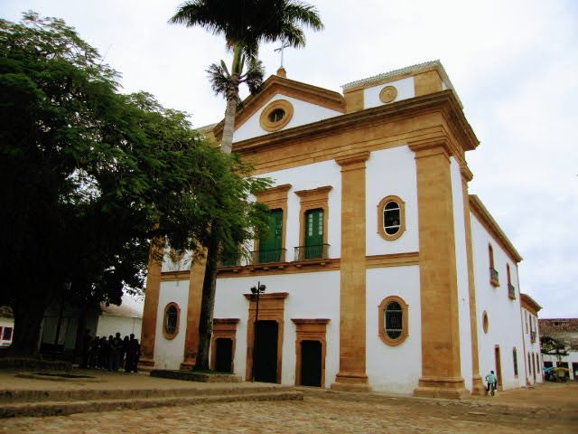 This image shows a colonial-style building with white walls and prominent beige or yellowish-brown columns framing its windows and doorways. The building appears to be a church, characterized by its symmetrical architecture, arched windows, and a central, triangular pediment at the top. There are green shutters on the windows and a palm tree in front, giving the setting a tropical feel. The building is situated on a cobblestone plaza, which adds to the historical atmosphere. The sky above is overcast, indicating a cloudy day.