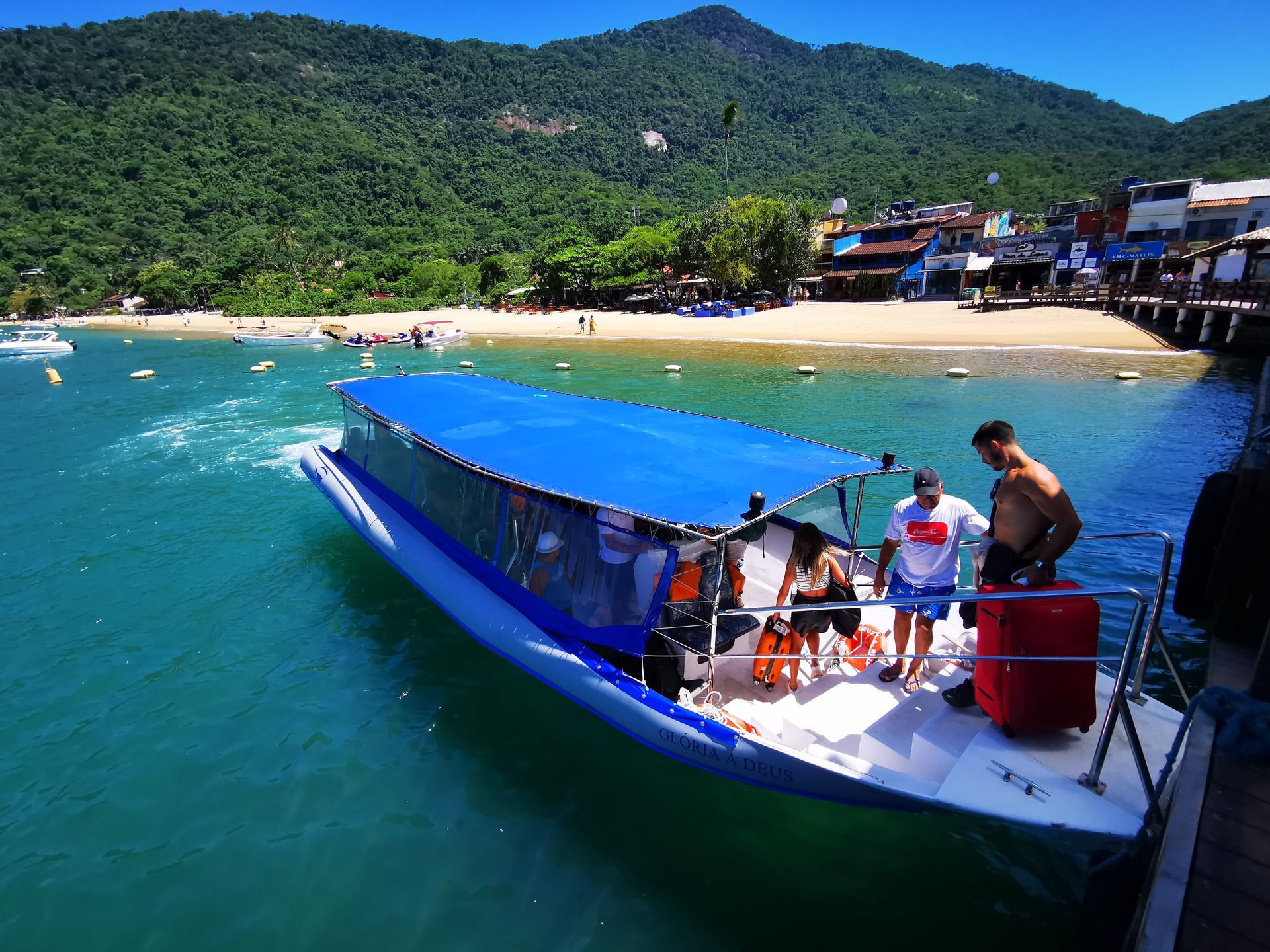 Barco com turistas saindo do píer da Vila do Abraão, em Ilha Grande, com águas calmas e cristalinas, casas à beira-mar e vegetação de Mata Atlântica ao fundo.