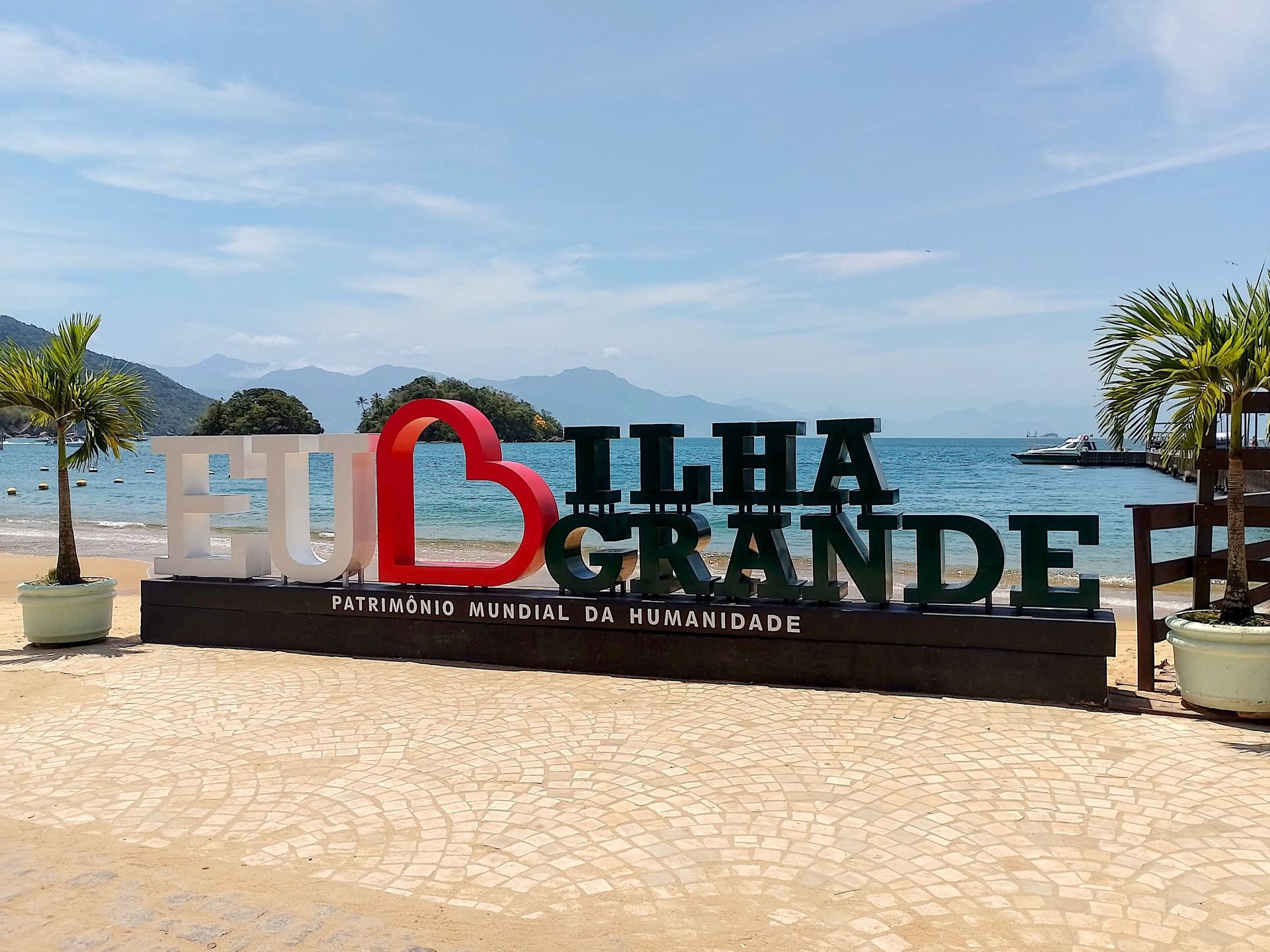  Eu Amo Ilha Grande” em frente à praia, com vista para o mar azul, ilhotas ao fundo e montanhas 