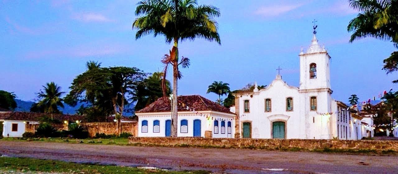 Igreja colonial e construções históricas em Paraty, Rio de Janeiro, com palmeiras e bandeirinhas coloridas ao entardecer.