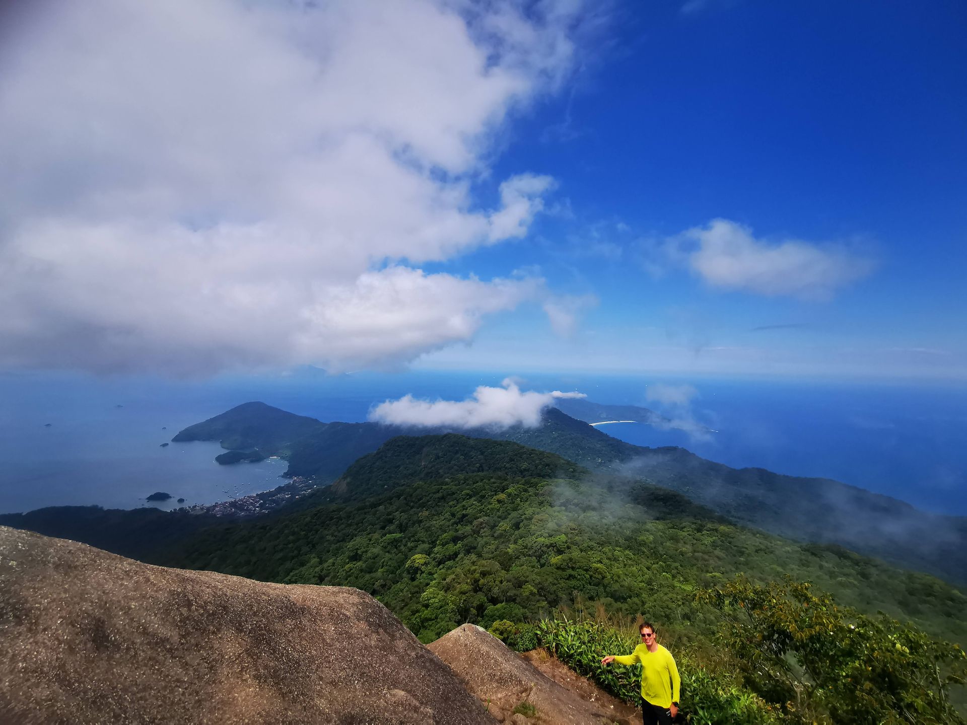 Hiker enjoying the panoramic view from Pico do Papagaio, Ilha Grande, with lush green mountains, ocean, and scattered clouds, capturing the beauty of this tropical island landscape