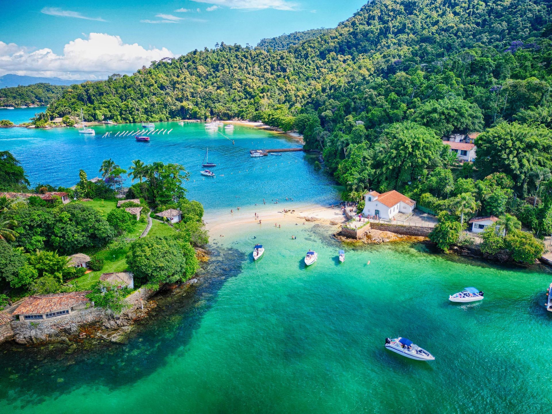 Aerial view of Praia da Piedade in Ilha Grande, Brazil, featuring crystal-clear waters, anchored boats, a historic chapel by the shore, and visitors enjoying the scenic tropical paradise