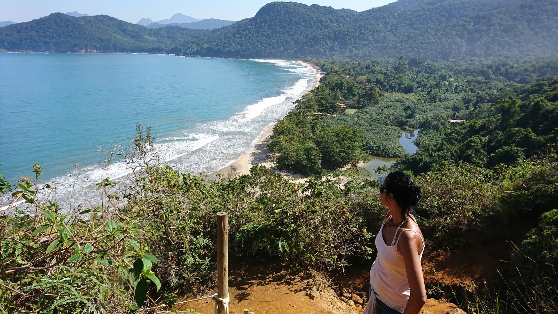  Woman standing on a cliffside trail overlooking Praia do Sono in Paraty, Brazil, with a panoramic view of the ocean, beach, forest, and surrounding mountains.