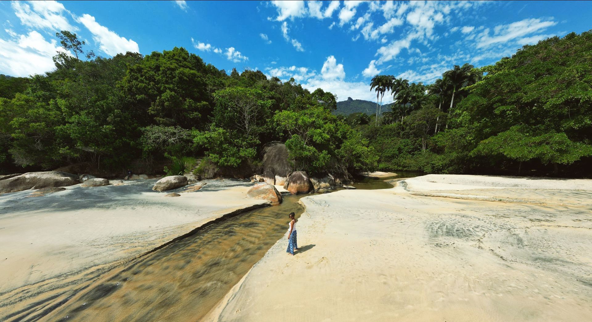 Tranquil scene at Praia Preta, Ilha Grande, featuring a person standing on the sandy beach near a small stream with lush green forest and large boulders in the background, showcasing the natural beauty and serene atmosphere of this unique Brazilian destination