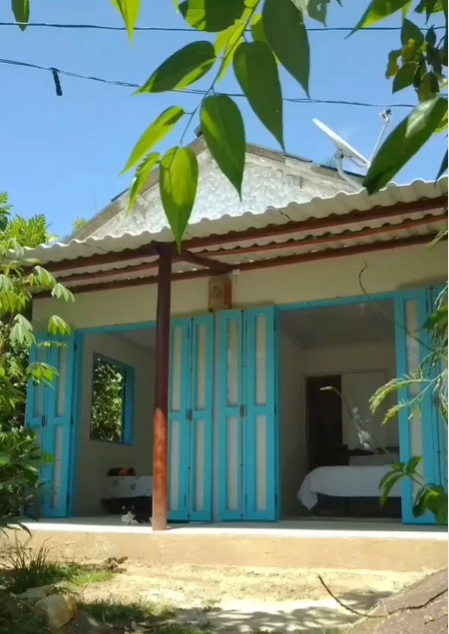 Vista externa de uma charmosa suíte no Varandinha em Aventureiro, Ilha Grande, com janelas de madeira azul abertas, cama aconchegante visível pela porta e cercada por folhagem verde exuberante, criando uma atmosfera serena e convidativa