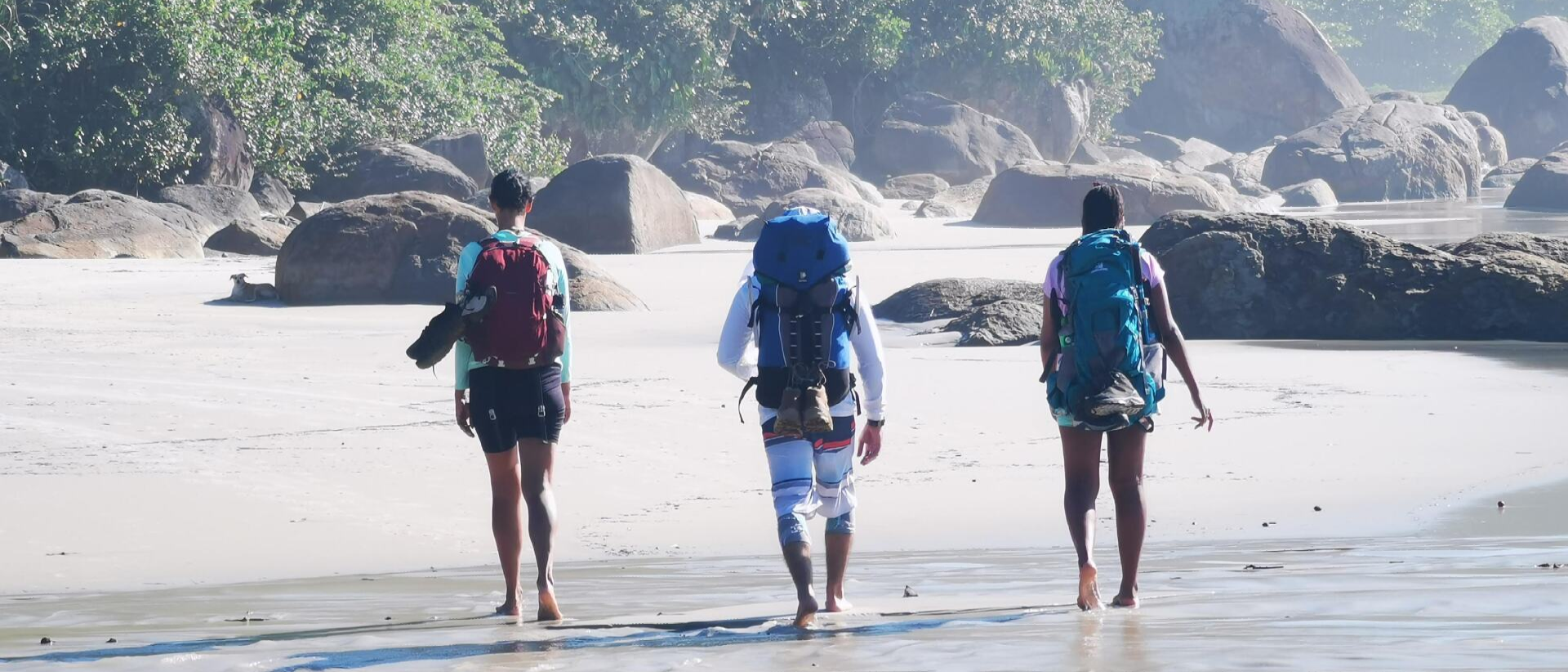 3 pessoas caminhando na praia com mochilas de caminhada.