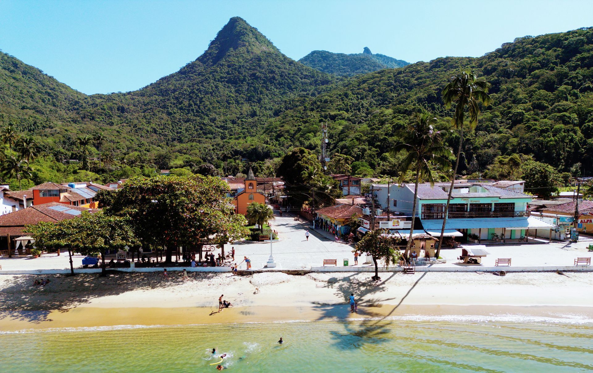 The image depicts Vila do Abraão, the main village on Ilha Grande. The beachfront shows calm, shallow waters with people enjoying the beach, while the village itself is nestled between lush, forested mountains. The buildings are colorful and quaint, reflecting the laid-back atmosphere of the island. Palm trees and greenery enhance the tropical feel of this popular destination, known for its natural beauty and tranquil surroundings. The towering peaks in the background, covered in dense vegetation, highlight the island's rugged landscape, offering a beautiful contrast to the serene beach.