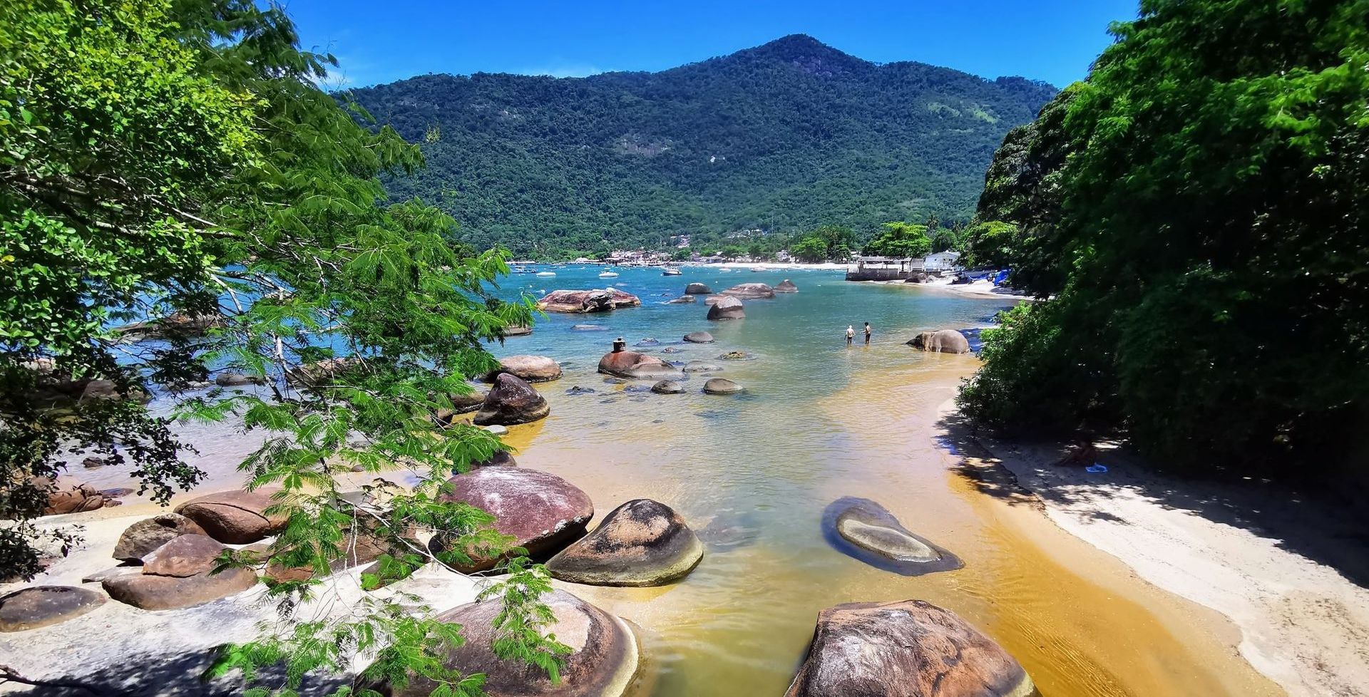 The image depicts Vila do Abraão, the main village on Ilha Grande. The beachfront shows calm, shallow waters with people enjoying the beach, while the village itself is nestled between lush, forested mountains. The buildings are colorful and quaint, reflecting the laid-back atmosphere of the island. Palm trees and greenery enhance the tropical feel of this popular destination, known for its natural beauty and tranquil surroundings. The towering peaks in the background, covered in dense vegetation, highlight the island's rugged landscape, offering a beautiful contrast to the serene beach.