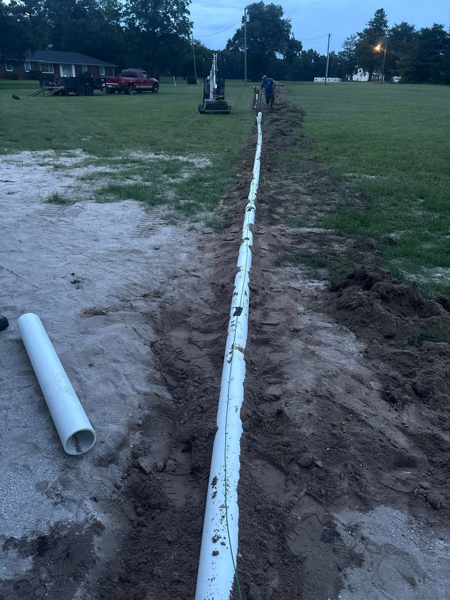 A long white PVC pipe is laid in a trench outdoors with workers in the background.