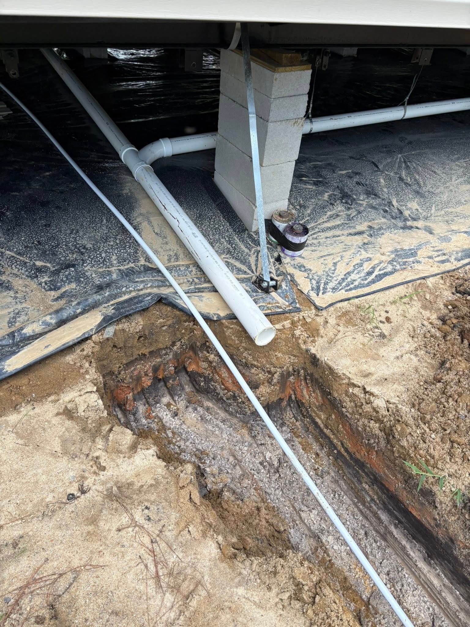 Underneath a house, a trench and gray pipes are visible. A white cable runs alongside them.