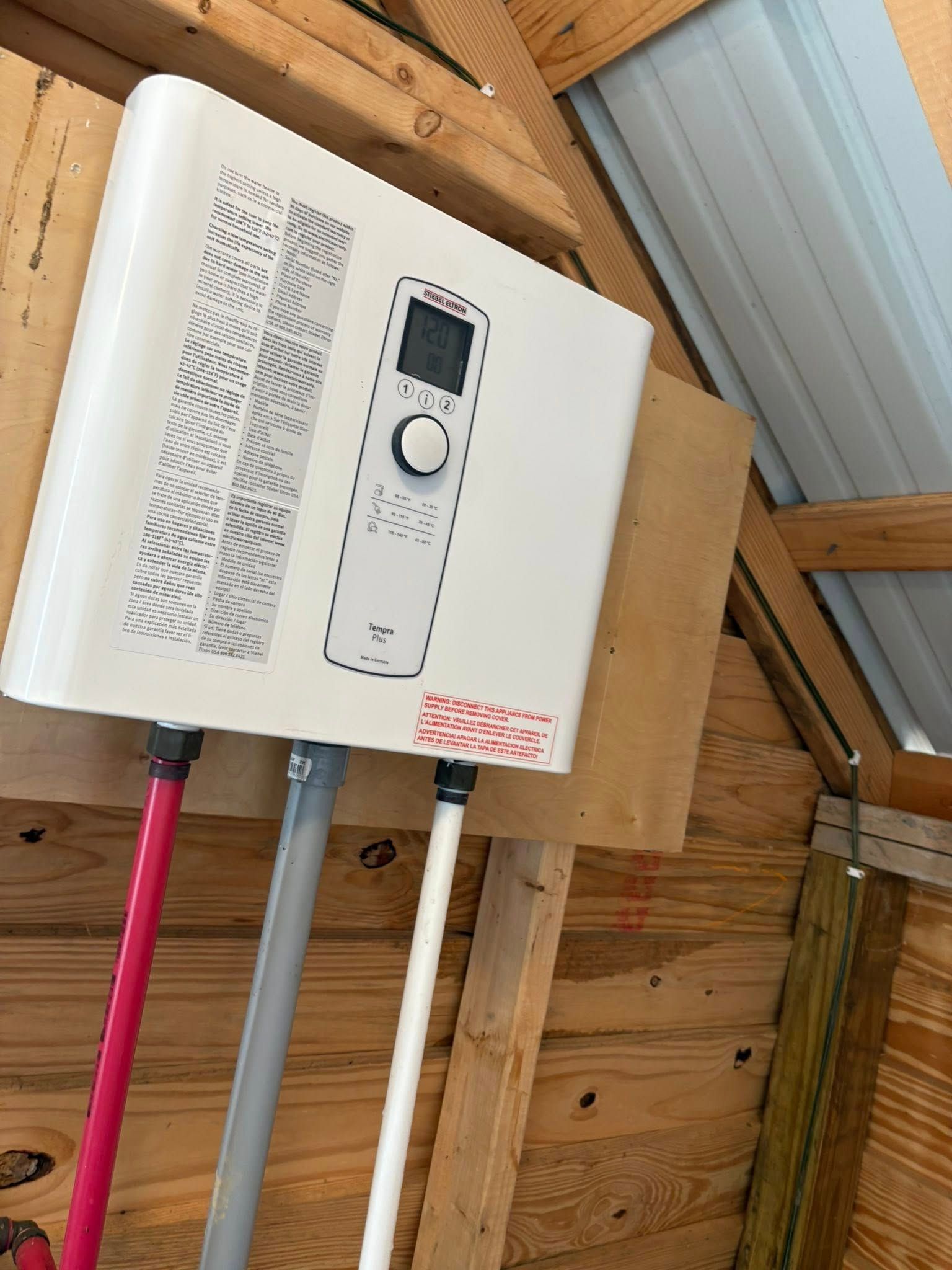 White tankless water heater mounted on a wooden wall, with red and white pipes.