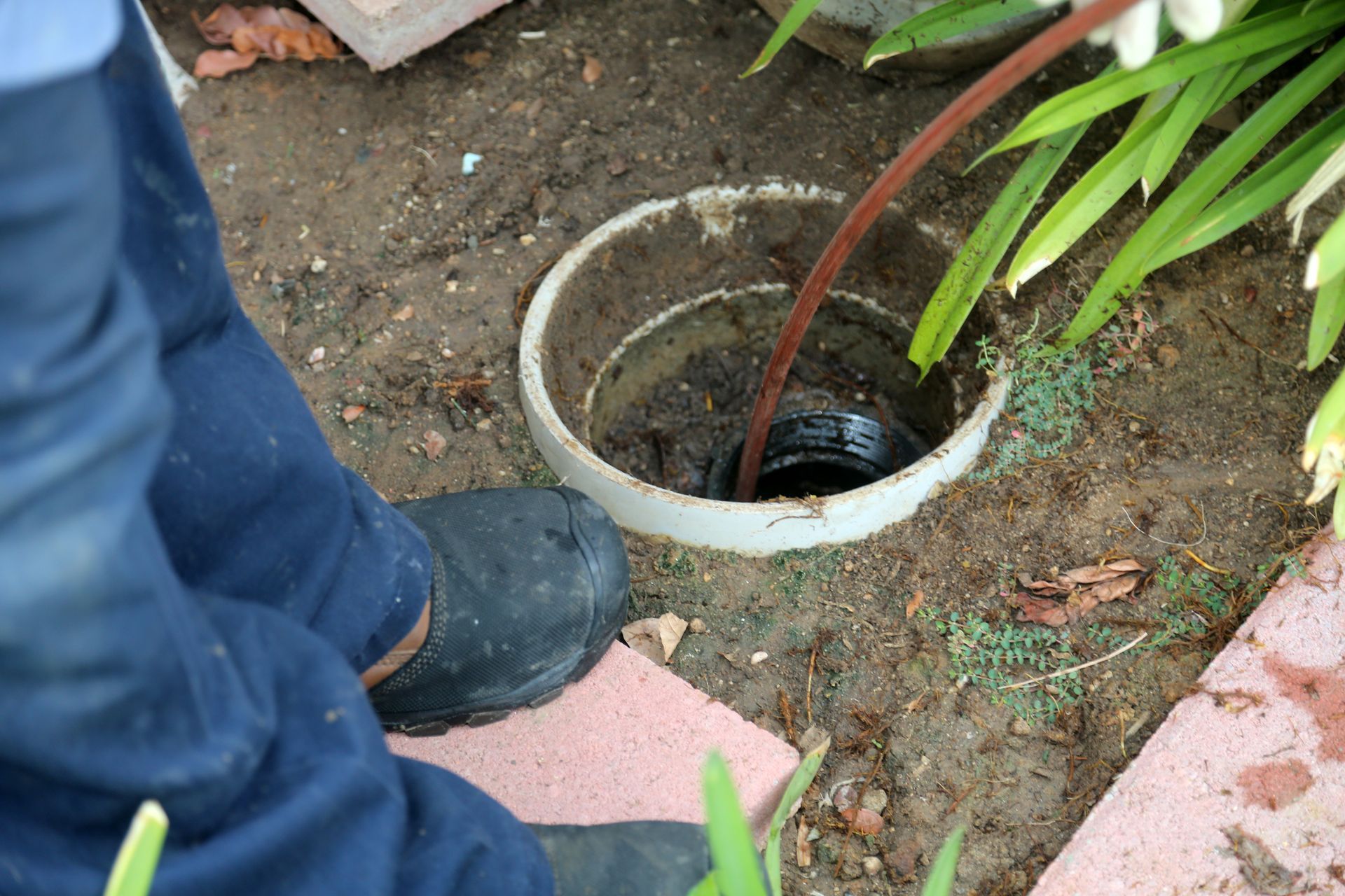 Person in blue pants and boots, using a hose to clear a drain in a dirt yard, next to flowers and a brick border.
