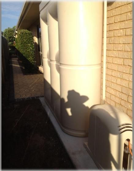 Tall Beige Water Tanks Next to A Brick Wall, with A Small Matching Unit at The Base — Sweeney's Pumps & Irrigation In Medowie, NSW