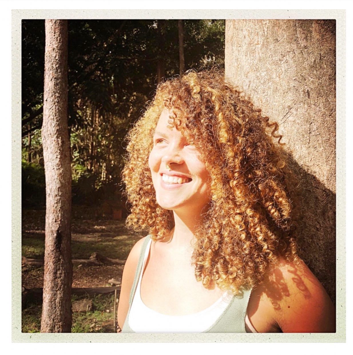 A woman with curly hair is smiling in front of a tree