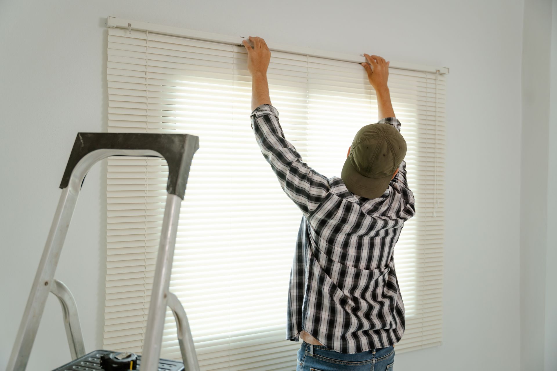 A professional blind installer adjusts sleek blinds in a modern house, standing next to a ladder.
