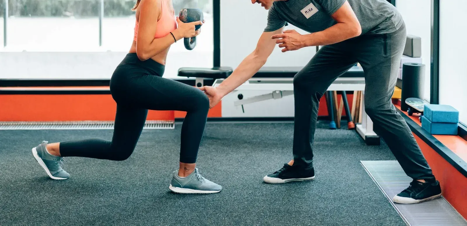 A man is helping a woman do lunges in a gym.