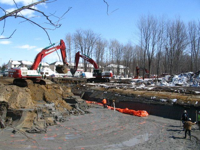 Construction site with excavators digging, surrounded by snow, trees, and buildings.