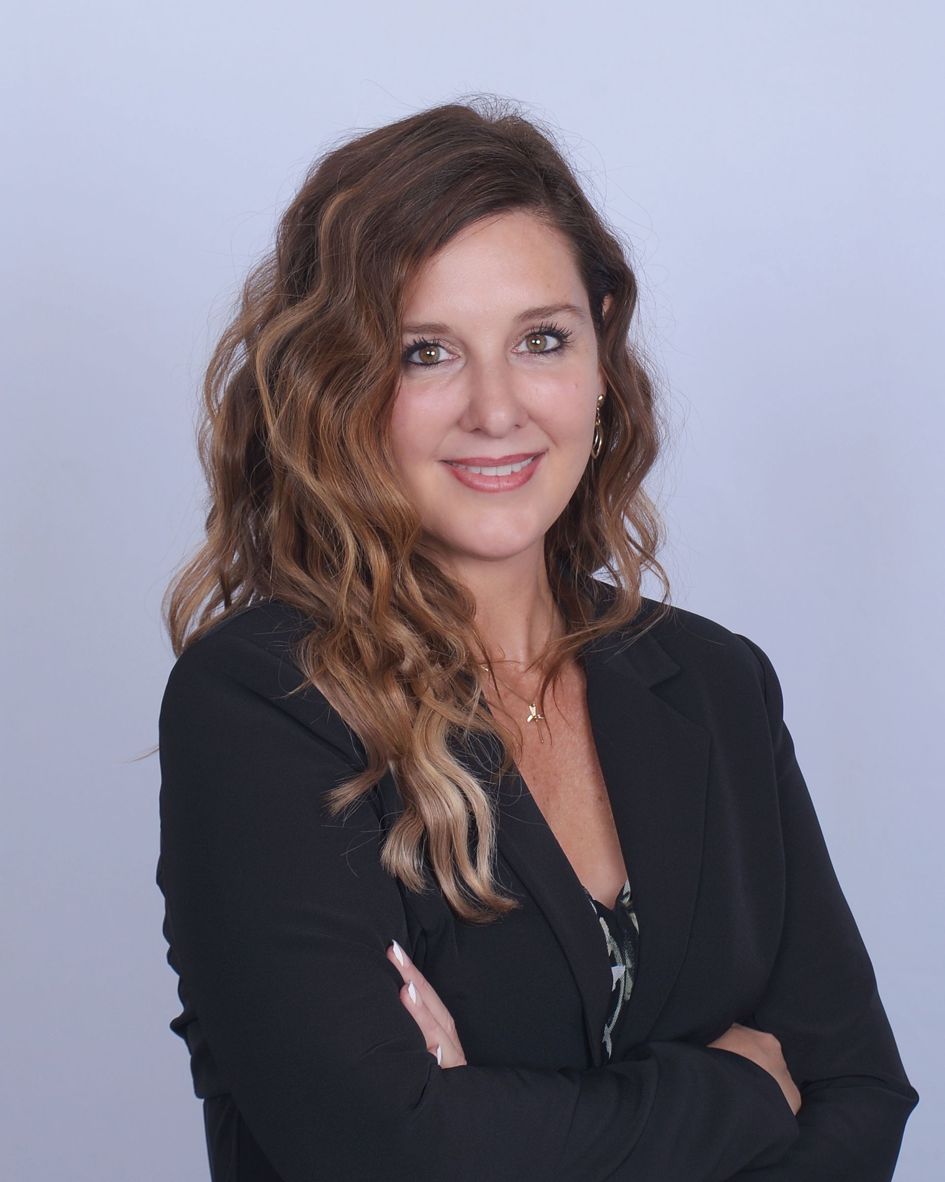 Portrait of a smiling woman with wavy hair in a black blazer, arms crossed, against a light blue background