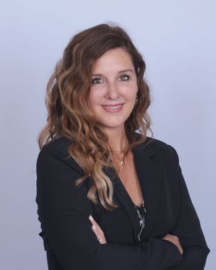 Portrait of a smiling woman with wavy hair in a black blazer, arms crossed, against a light blue background