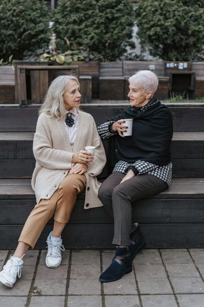 Two women sit on steps outdoors, talking over drinks in a park-like setting.